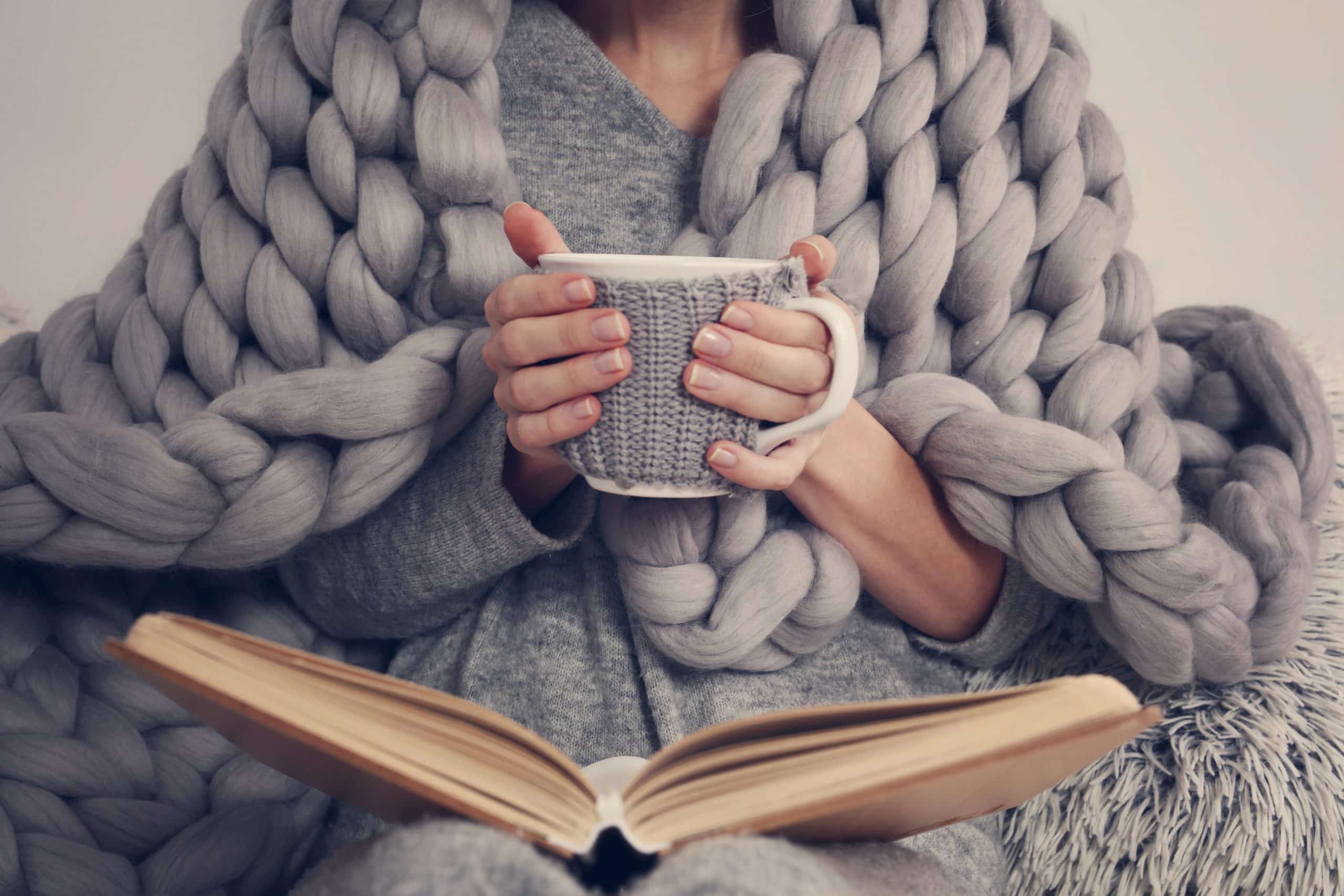 woman in weighted blanket holding mug and reading book in lap